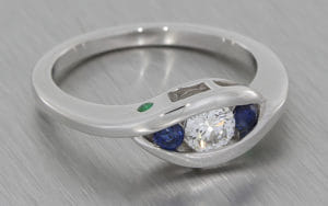 Bypass style white gold trilogy ring set with a a round brilliant diamond with two round sapphires either side